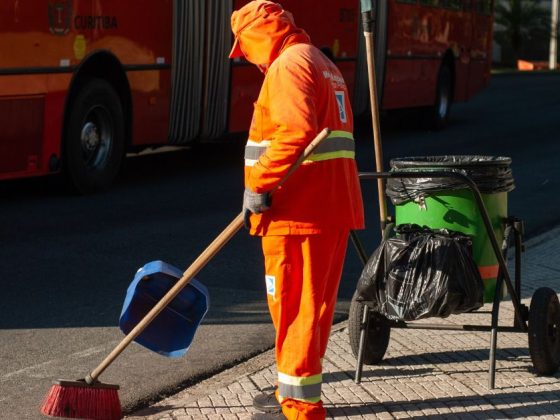 Varredora de rua com uniforme laranja varre calçada ao lado de carrinho de lixo — direitos trabalhistas