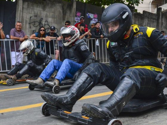 Competidores com capacete e equipamentos de proteção disputam corrida de carrinho de rolimã em rua com público ao fundo