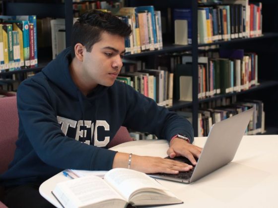 Estudante usando notebook em biblioteca com livro aberto, representando o uso da rede social para concurseiros
