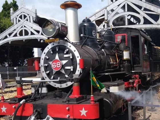 Maria Fumaça, locomotiva a vapor número 68, parada na estação com vapor saindo da parte frontal e bandeira do Brasil na grade