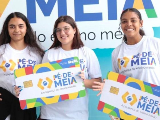 Três jovens estudantes sorrindo lado a lado, vestindo camisetas brancas do programa Pé-de-Meia, seguram cartões do benefício em frente a um painel com o logo "Pé-de-Meia: a poupança do ensino médio"