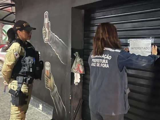 Agente da fiscalização da Prefeitura de Juiz de Fora afixa lacre de interdição no bar interditado em Juiz de Fora enquanto policial militar acompanha a ação