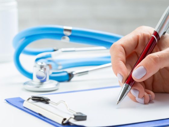 Mão de profissional preenchendo atestado médico que causou demissão por justa causa, ao lado de estojo de comprimidos e estetoscópio azul