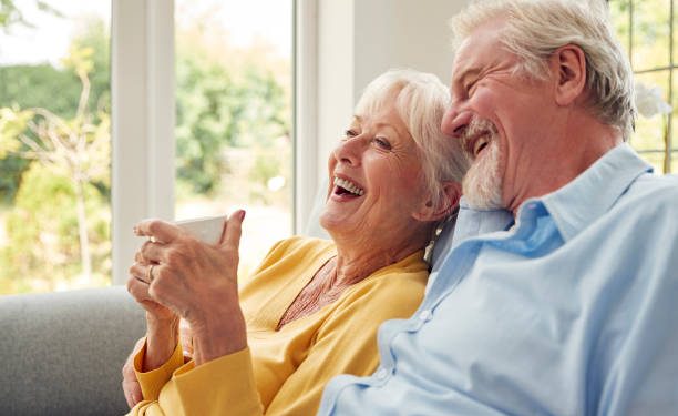 Retired Senior Couple Sitting On Sofa At Home Drinking Coffee And Watching TV Together