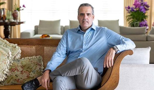 um homem está sentado com uma perna cruzada sob a outra e segurando esta com uma mão, ele usa camisa social azul clara, uma calça bege e tenis preto