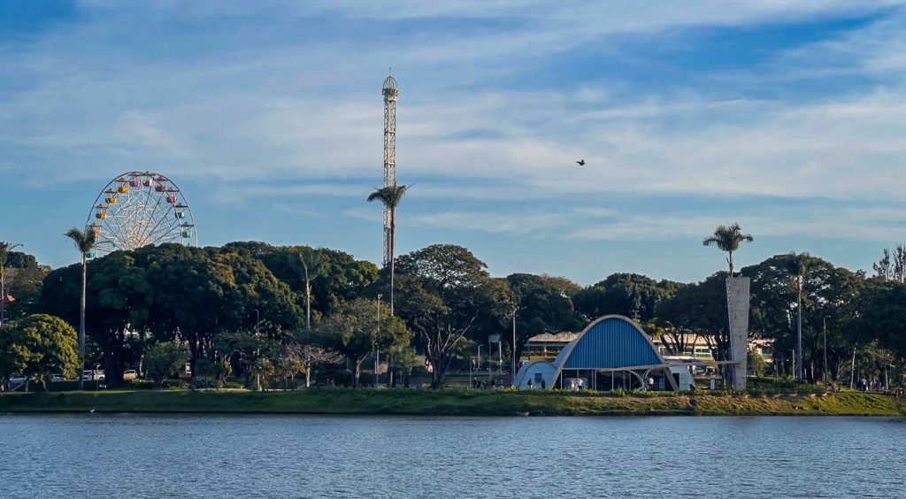 Foto panorâmica da Lagoa da Pampulha com a Igrejinha ao fundo e a roda gigante de um parque de diversões.