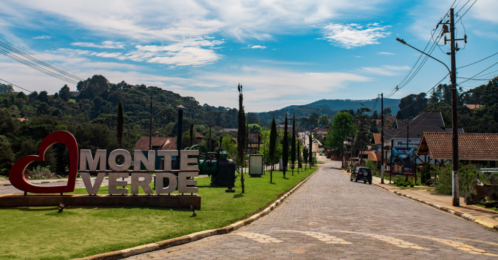 Paisagem de Monte Verde, em Minas Gerais, conhecida por suas altas temperaturas. Imagem mostra cenário da cidade, com casas, morros e caminho de pedras, e placa com o nome da cidade. 