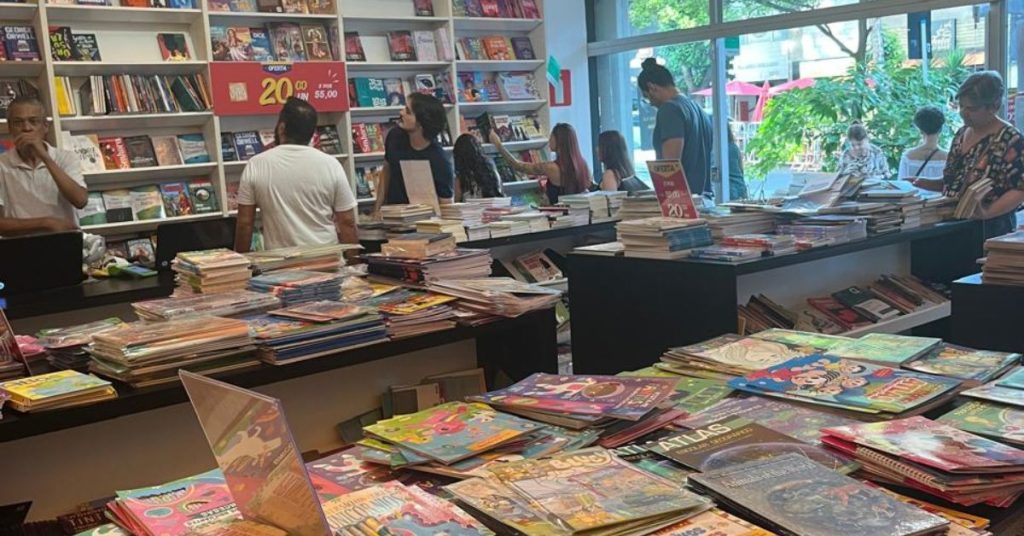 Imagem interna da livraria Outlet de Livro mostra várias opções de livros nas prateleiras e estandes, e clientes circulando pelo local.