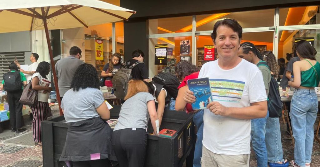 O empresário José Henrique Guimarães, dono do Outlet de Livros, mostra um dos títulos a R$ 1 em sua loja, e várias pessoas atrás dele mexem nos livros nas bancas dispostas na área externa.