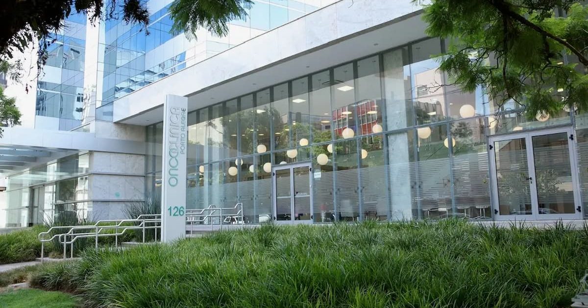 Fachada de um edifício comercial moderno, com ampla fachada de vidro e estrutura branca. Na entrada, há portas de vidro, áreas ajardinadas com gramado e árvores, além de mesas e luminárias visíveis no interior do prédio. Na lateral da fachada, uma placa vertical exibe o nome do edifício.