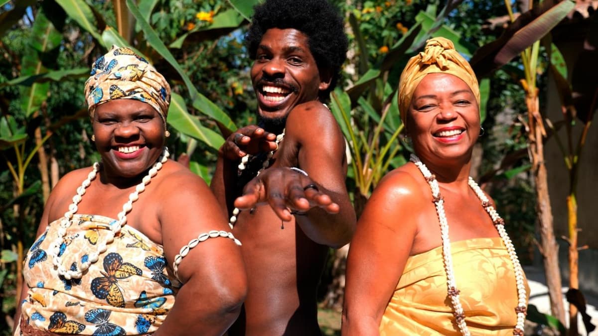 Três pessoas afro-brasileiras sorrindo, duas mulheres com turbantes e um homem com cabelo afro, em um cenário verde tropical.