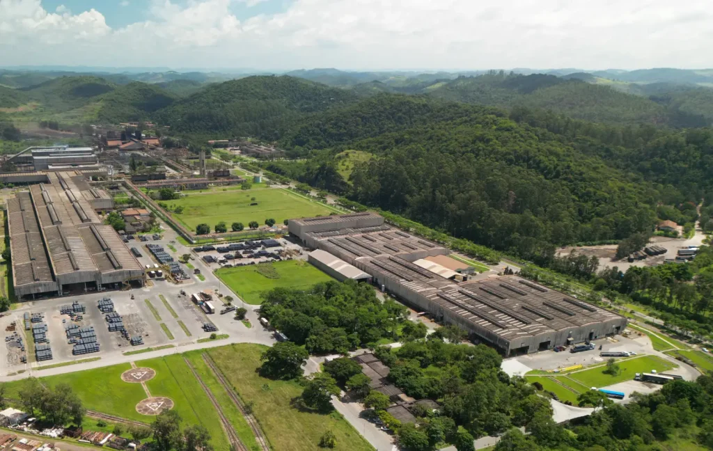 Vista aérea de um grande complexo industrial com extensos galpões e pátios de estocagem de materiais, cercado por colinas verdes e florestas sob um céu azul claro
