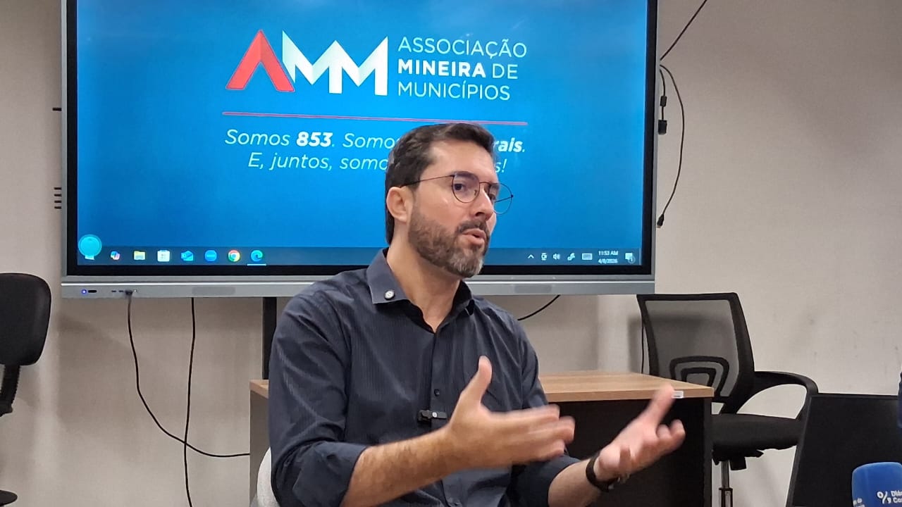 Homem com óculos e barba gesticulando em frente a um telão com o logo da Associação Mineira de Municípios (AMM).