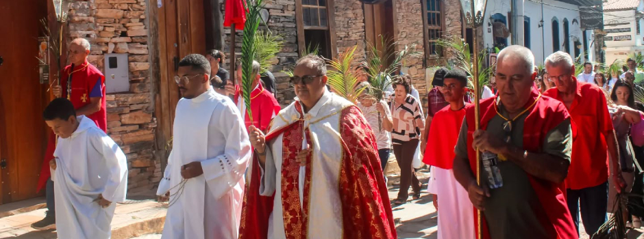 Procissão religiosa com padre e fiéis em rua histórica, carregando ramos de palmeira.