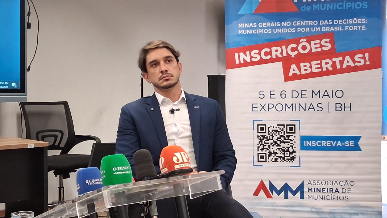 Homem de terno em coletiva de imprensa com banner da AMM ao fundo.