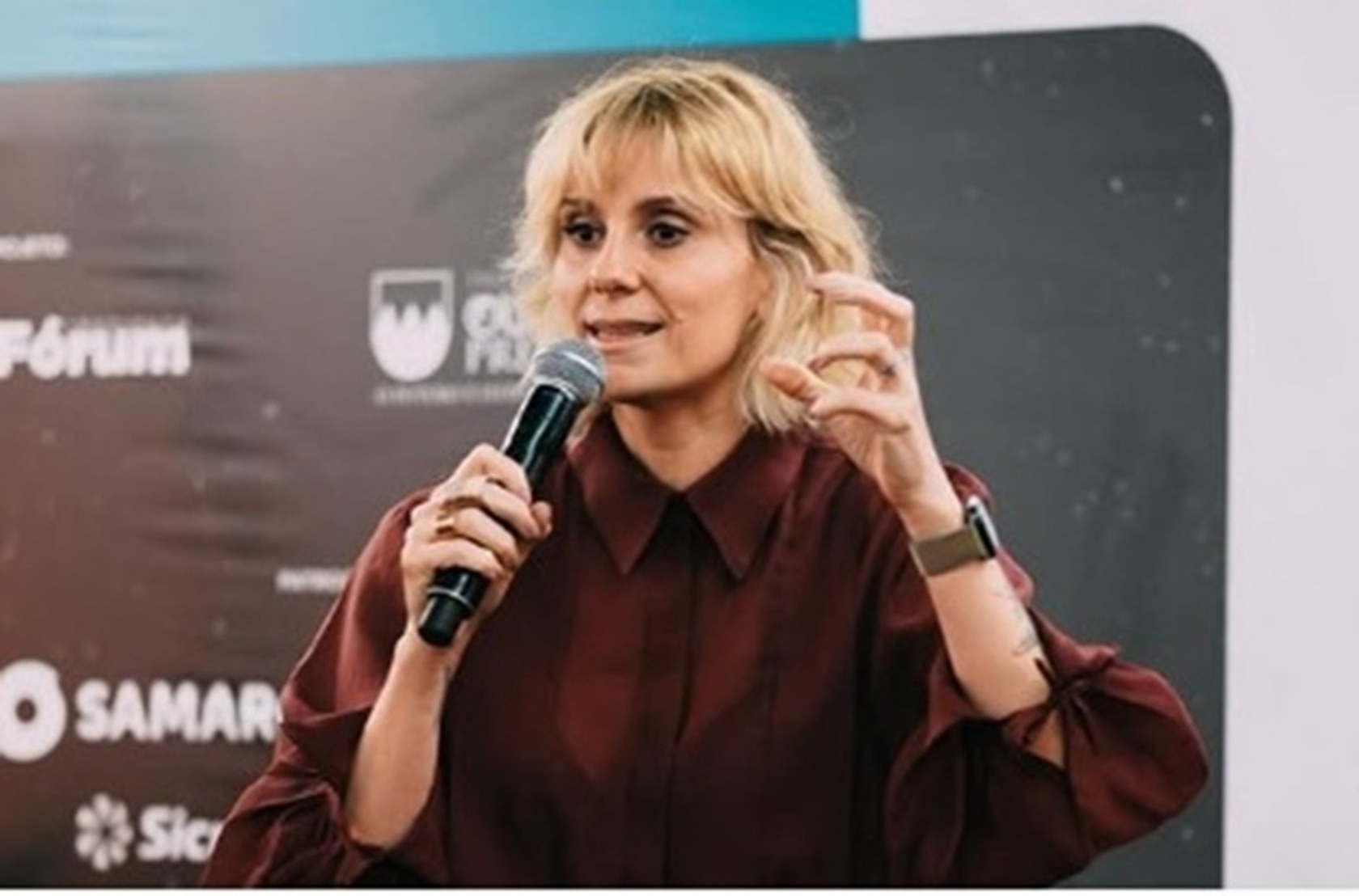 Mulher loira discursando com microfone em evento.