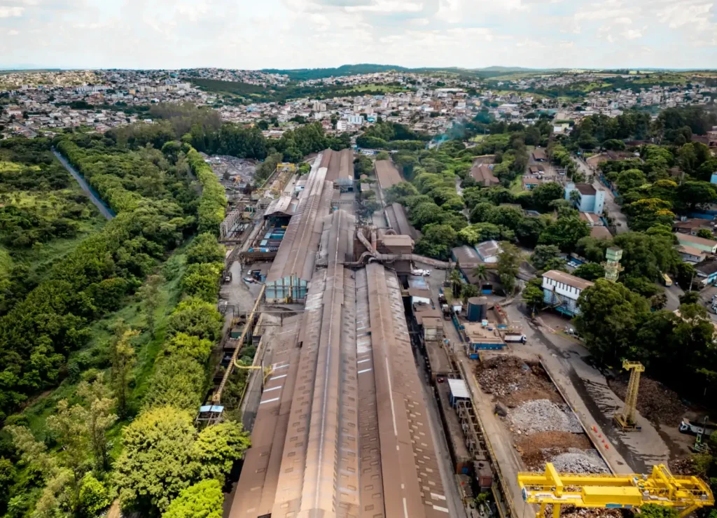 Fotografia aérea de um vasto complexo industrial com longos galpões metálicos e pátios de materiais, ladeado por uma densa vegetação e com uma cidade ao fundo sob um céu nublado.