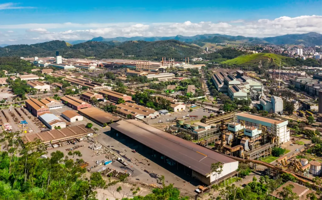 Vista aérea panorâmica de um grande complexo industrial, com diversos galpões, chaminés e infraestrutura de produção, em um vale rodeado por colinas verdes.