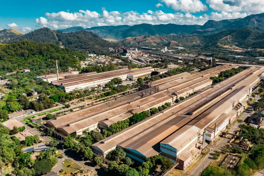 Vista aérea panorâmica de uma grande planta industrial com longos galpões e infraestrutura de transporte, cercada por exuberantes montanhas verdes sob um céu azul e nublado.