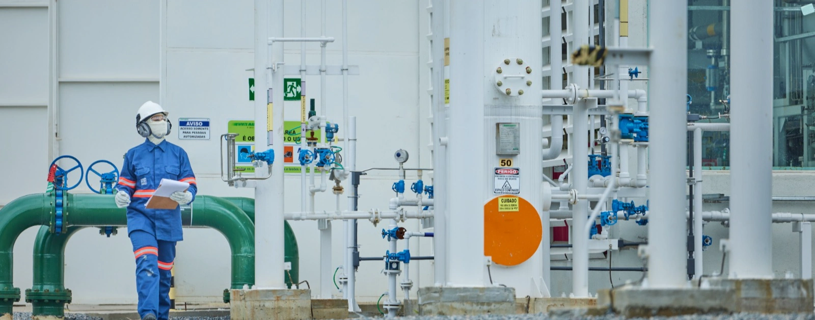 Pessoa em uniforme industrial azul e capacete branco caminhando em planta.