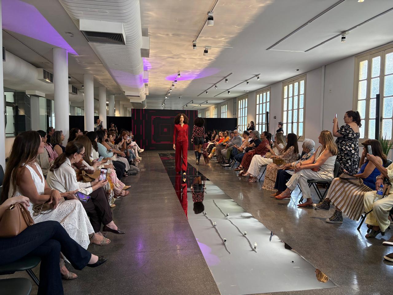 Desfile de moda em ambiente interno com mulher na passarela e espectadores em cadeiras.