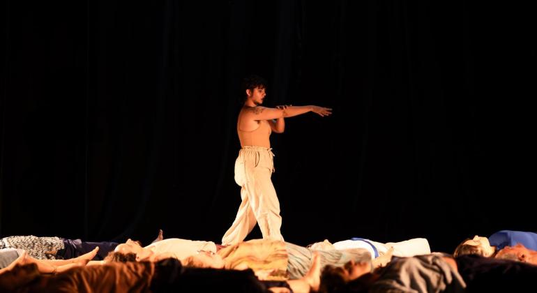 Mulher em pose de movimento em palco escuro, com pessoas deitadas no chão à sua frente.