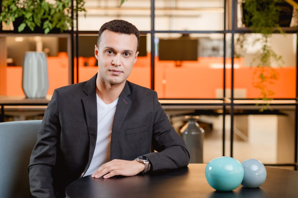 Homem jovem com blazer preto e camiseta branca sentado em mesa de escritório.