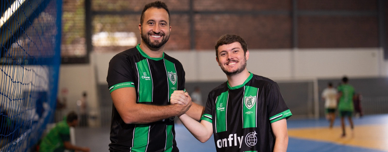Esta fotografia captura um momento de conexão e parceria entre dois indivíduos em um ambiente esportivo. Ambos estão trajados com uniformes distintivos nas cores verde e preta, trocando um aperto de mão e olhares amigáveis diretamente para a câmera.