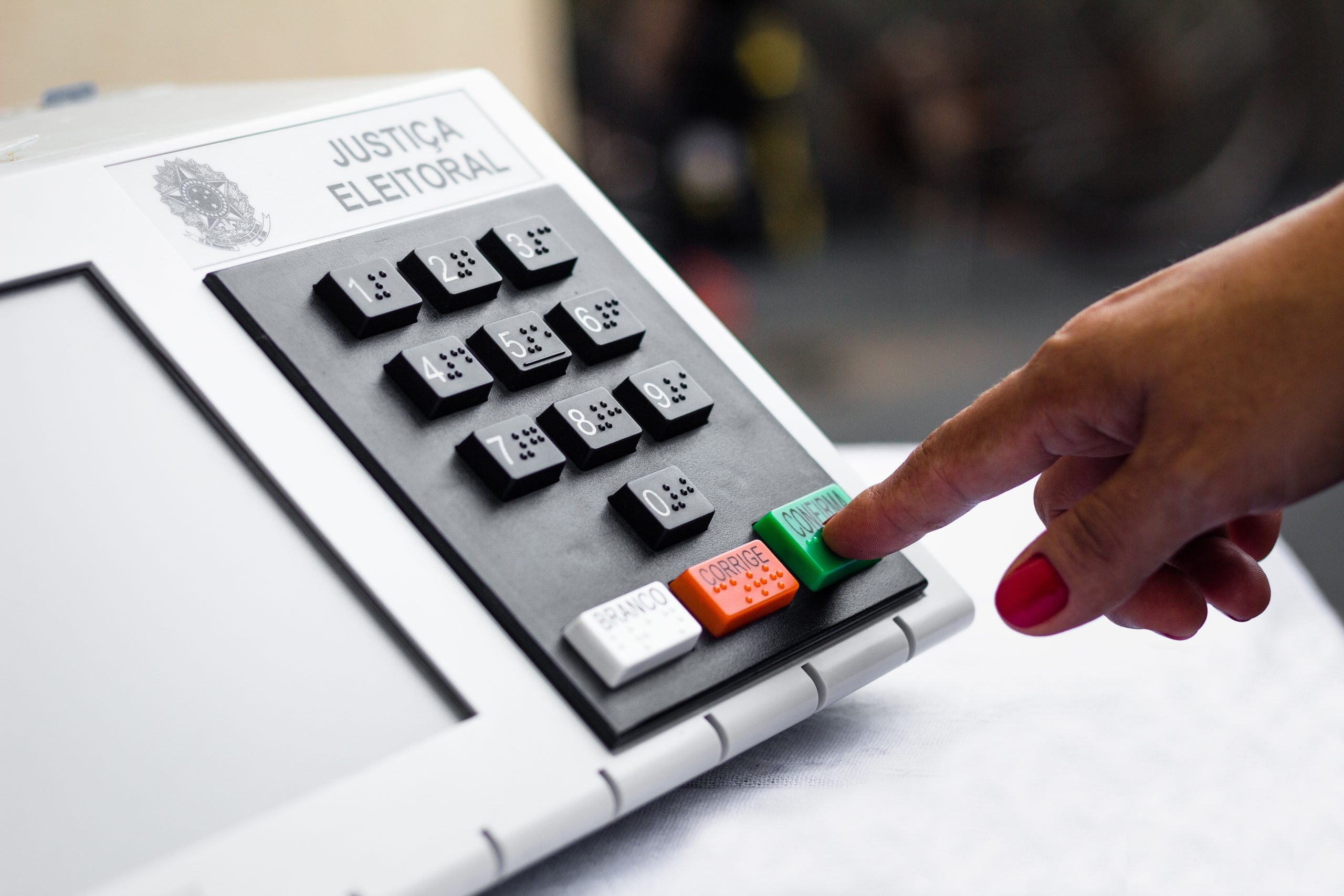 Mão apertando o botão verde 'Confirma' em urna eletrônica da Justiça Eleitoral.