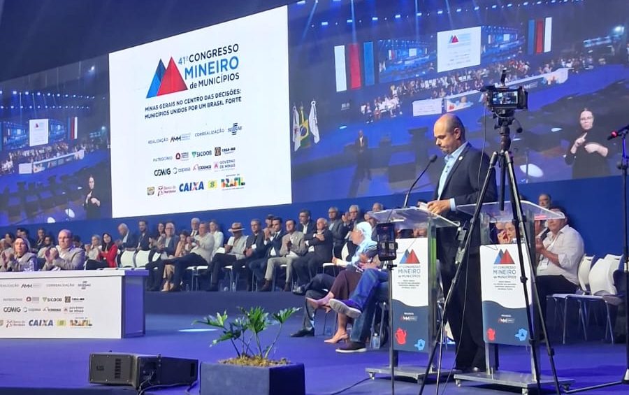 Um homem em terno escuro discursa com entusiasmo em um púlpito no palco de um grande evento. Ao fundo, uma tela de LED exibe o logotipo de um congresso