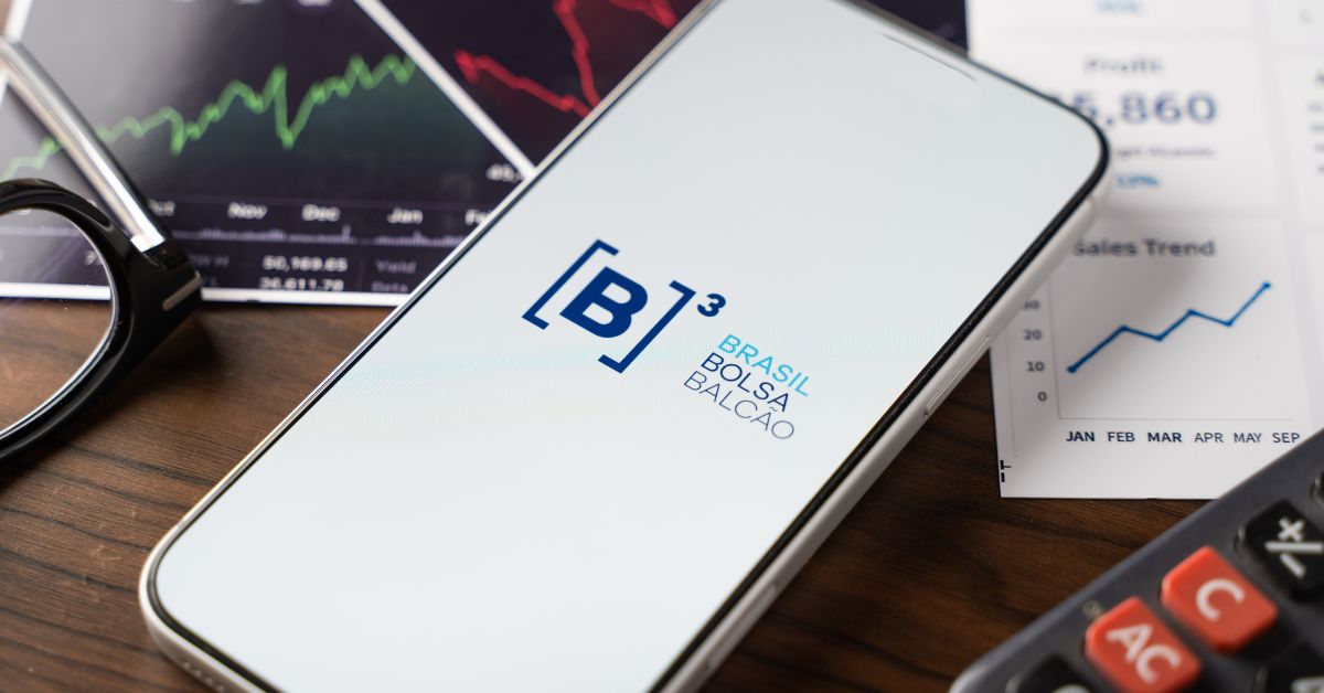 Aparelho celular exibe o logo da B3 sobre mesa com gráficos financeiros, calculadora e documentos, em referência às ferramentas digitais usadas por investidores na declaração do Imposto de Renda 2026.
