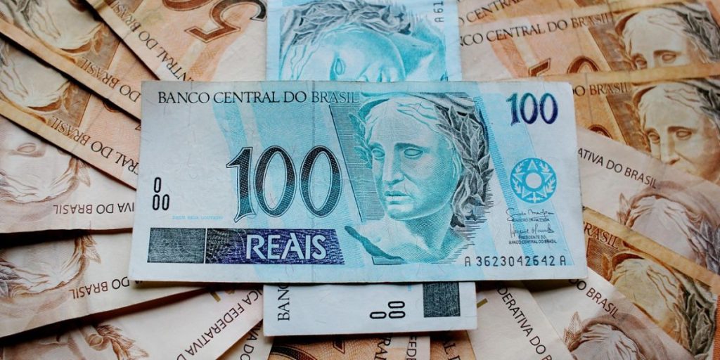 Notas de R$ 100 espalhadas ao fundo destacam uma cédula em primeiro plano. A imagem representa dinheiro em espécie, finanças, economia e poder de compra no Brasil.