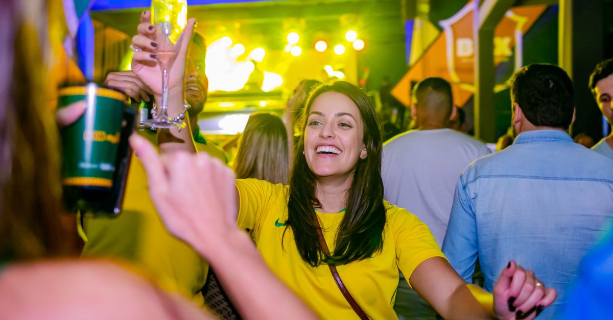 Mulher sorridente com camisa amarela em festa noturna e multidão ao fundo.