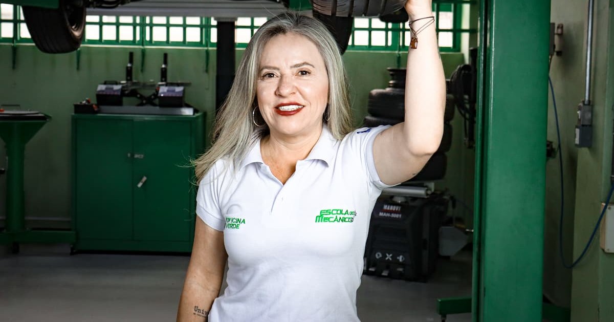 Mulher loira sorrindo em uma oficina mecânica vestindo camisa polo branca.