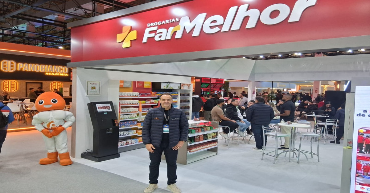 Homem de tênis branco, calça e jaqueta preta, posa ao lado de um stand de farmácia.