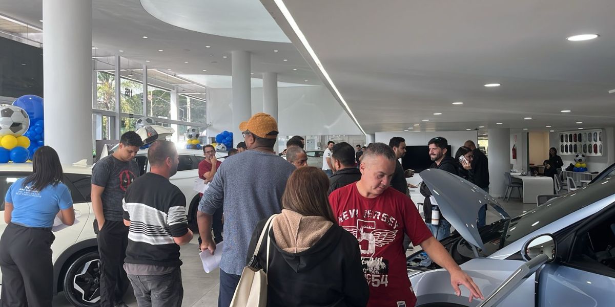 Interior de uma concessionária de veículos movimentada, com diversas pessoas observando e conversando sobre automóveis expostos. Em primeiro plano, um homem de camiseta vermelha analisa um carro prata com o capô aberto. Ao fundo, clientes circulam pelo showroom, que possui iluminação moderna, grandes colunas brancas e decoração com balões azuis, amarelos e bolas de futebol. Alguns veículos estão expostos para avaliação e negociação.
