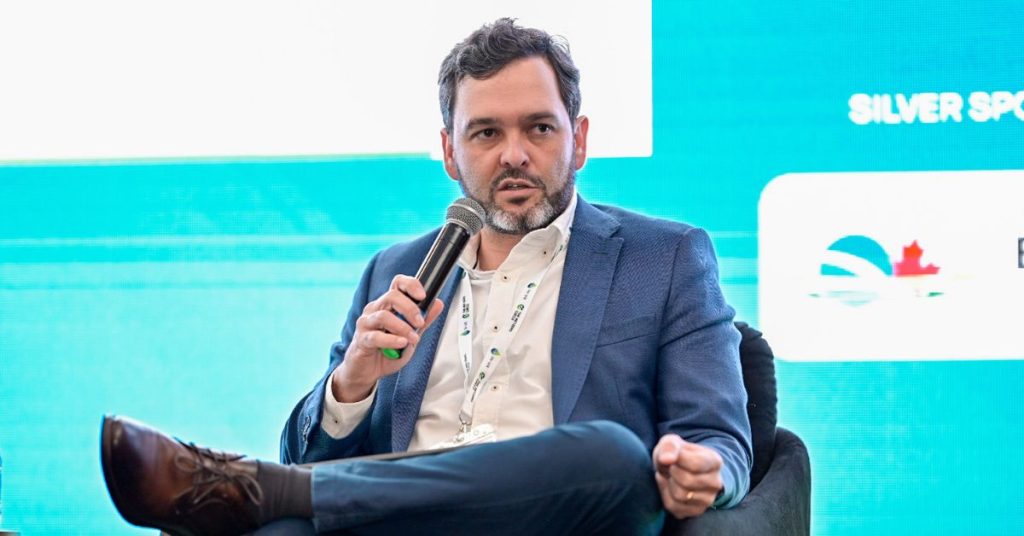 Ricardo Alves, Diretor Executivo da Graphcoa, fala em um painel de discussão em uma conferência, segurando um microfone e com uma perna cruzada. Ele usa um blazer azul, camisa branca e cordão de credencial. O fundo é uma tela LED com o texto "SILVER SPONSOR" e logotipos de patrocinadores.