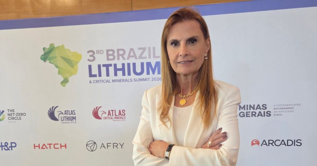 Uma foto de Marisa Cesar, Diretora de Assuntos Corporativos e Sustentabilidade da PLS, posando em um evento corporativo. Ela está em pé, com os braços cruzados, sorrindo para a câmera. Atrás dela, há um banner de fundo que diz "3rd BRAZIL LITHIUM & CRITICAL MINERALS SUMMIT 2026" com logos de empresas parceiras. A foto é cortada acima da cintura.