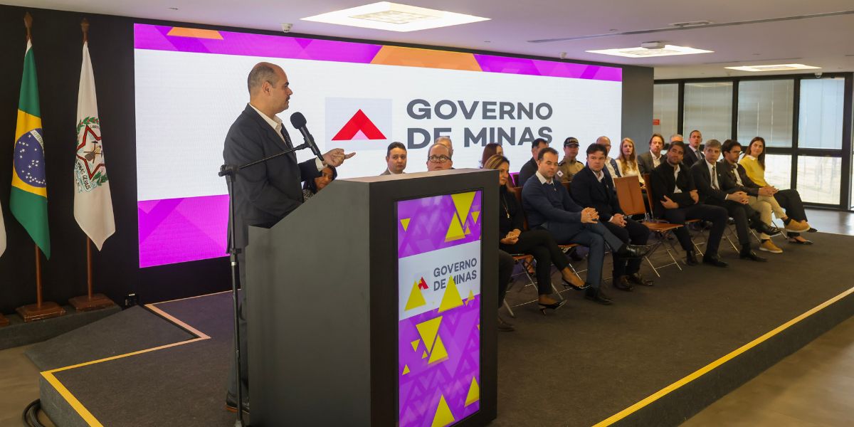 Autoridade do Governo de Minas discursa em cerimônia de assinatura de contrato, em auditório. Ao fundo, um telão exibe a logomarca do Governo de Minas, enquanto autoridades e convidados acompanham o evento sentados no palco. Bandeiras do Brasil e de Minas Gerais aparecem à esquerda da imagem.