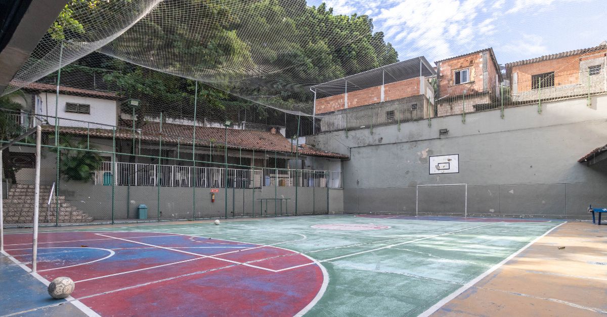 Quadra poliesportiva cercada por telas de proteção, com marcações para futsal e basquete em piso colorido. Ao fundo, há um prédio de apoio com varanda coberta e, acima do muro que delimita o espaço, aparecem casas em área de encosta com vegetação densa. O local está vazio e iluminado pela luz do dia.