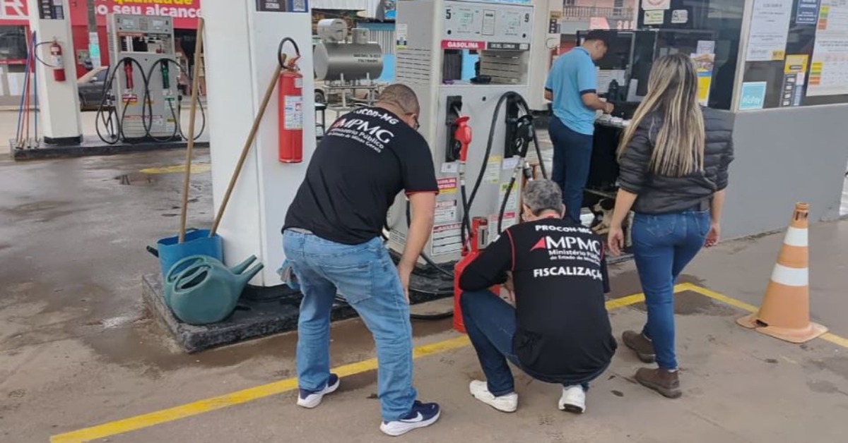 Fotografia mostra uma ação de fiscalização em um posto de combustíveis. Em primeiro plano, três fiscais do Procon-MPMG (Ministério Público de Minas Gerais) inspecionam uma bomba de combustível. Dois deles usam camisetas pretas com a identificação do órgão nas costas; um está agachado observando a parte inferior da bomba, enquanto o outro se inclina para acompanhar a vistoria. Ao lado, uma mulher de cabelos longos, vestindo jaqueta preta e calça jeans, acompanha o trabalho.