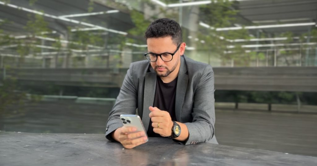 Homem adulto de cabelos curtos e escuros está sentado à mesa em um ambiente corporativo moderno. Ele veste blazer cinza sobre camiseta preta, usa óculos de armação escura e relógio no pulso esquerdo. Com expressão concentrada, segura um smartphone com uma das mãos enquanto observa atentamente a tela.