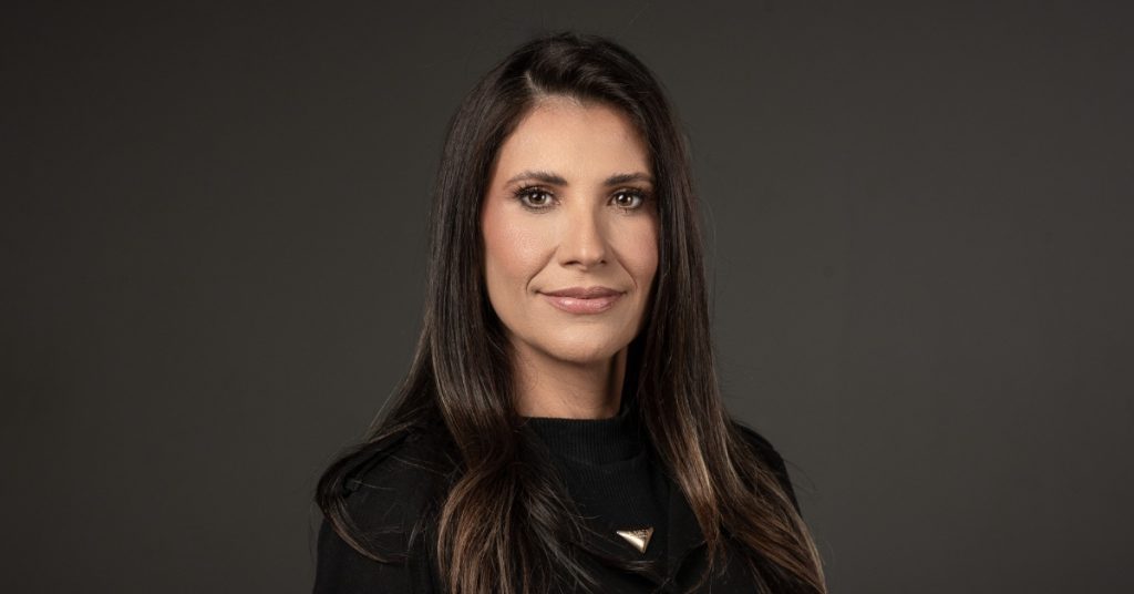 Retrato profissional de uma mulher adulta, de pele clara e cabelos longos castanho-escuros, levemente ondulados nas pontas. Ela está posicionada de frente para a câmera, com expressão serena e discreto sorriso. Usa maquiagem suave, destacando os olhos e os lábios em tom neutro. Veste uma blusa preta de gola alta e um acessório metálico dourado em formato triangular próximo ao pescoço.