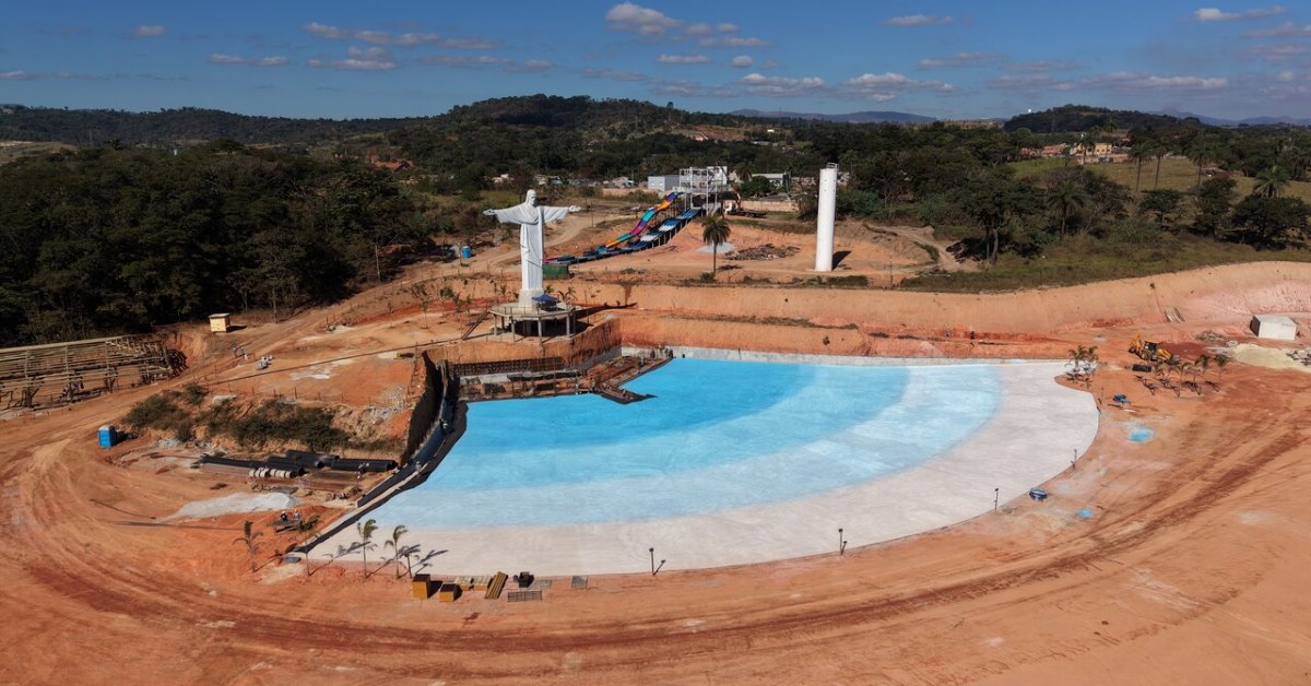 Fotografia aérea de uma grande obra em fase de construção, cercada por áreas de terra avermelhada e vegetação. No centro da imagem destaca-se uma ampla piscina artificial de formato irregular, com revestimento azul-claro que lembra uma praia de águas rasas. À esquerda da piscina há uma estátua branca inspirada no Cristo Redentor, com os braços abertos sobre uma plataforma elevada.