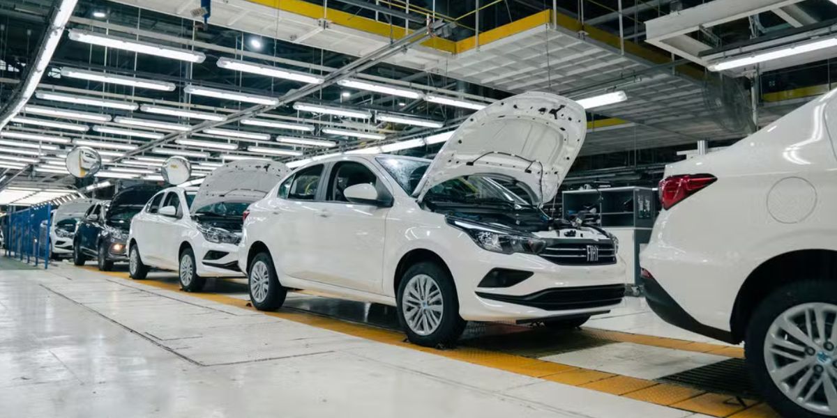 Linha de produção da fábrica da Stellantis em Betim (MG), com veículos Fiat brancos em processo de montagem e inspeção, posicionados em sequência dentro da unidade industrial, sob iluminação e estrutura fabril.