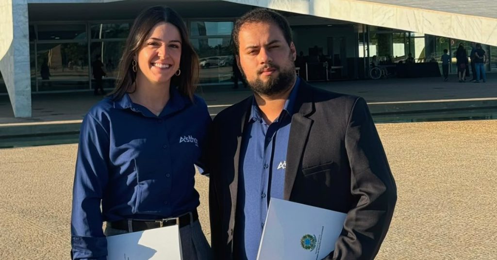 Duas pessoas posam lado a lado em frente a um edifício moderno de arquitetura monumental, com grandes colunas e fachada envidraçada. A cena parece ter sido registrada durante o dia, sob luz solar intensa.
À esquerda está uma mulher de cabelos longos e escuros, sorrindo para a câmera. Ela usa camisa social azul-marinho com o logotipo de uma empresa bordado no peito, brincos de argola e segura uma pasta ou documento branco.
À direita está um homem de barba e cabelos curtos escuros. Ele veste camisa azul-marinho semelhante à da mulher, com o mesmo logotipo, além de um blazer escuro. Também segura uma pasta branca, na qual aparece o brasão da República Federativa do Brasil.