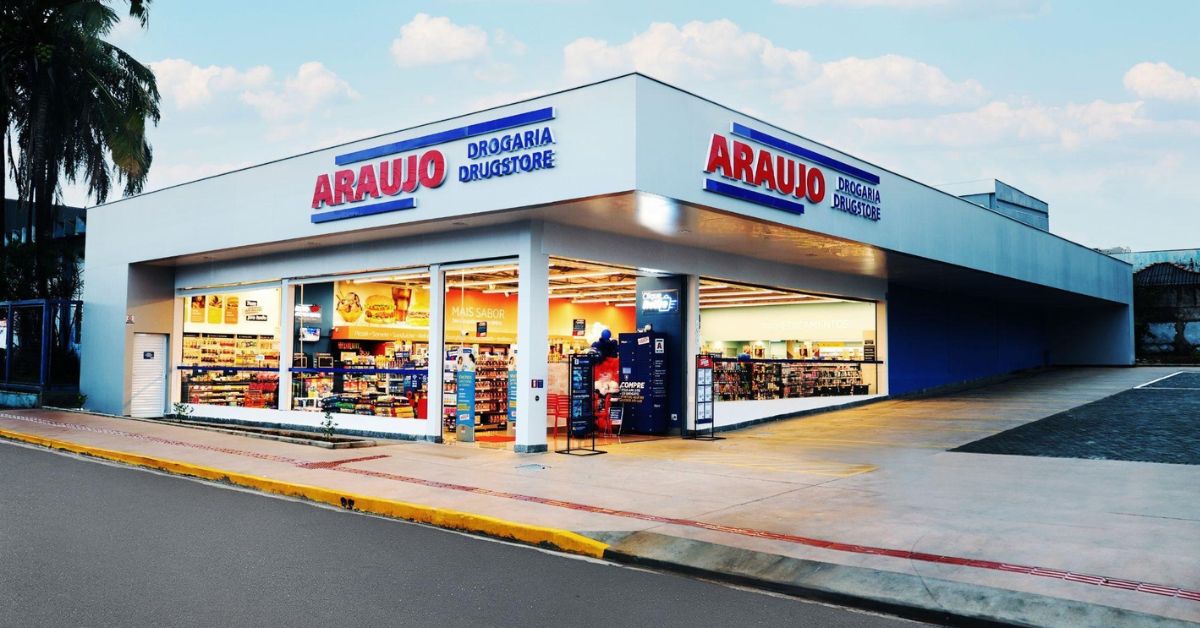 Fachada de uma unidade da Drogaria Araujo em Uberaba. A loja, de esquina, tem fachada branca com letreiros em vermelho e azul, amplas vitrines de vidro que permitem ver o interior iluminado e estacionamento ao lado. A imagem foi registrada ao entardecer, com céu parcialmente nublado.
