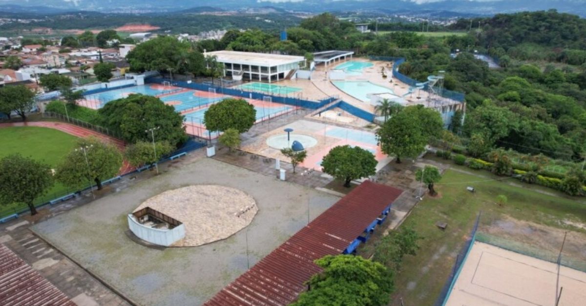Vista aérea do antigo Clube da Fiat, em Betim, mostrando ampla área com piscinas, quadras esportivas, campo de futebol, edifícios e árvores. O espaço, cercado por vegetação e áreas urbanas ao fundo, será transformado em campus da UFMG.