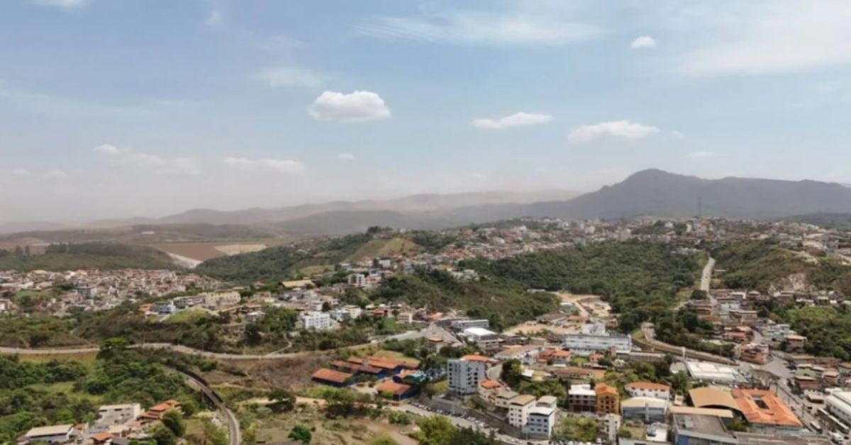 Vista panorâmica de Congonhas, na região Central de Minas Gerais, com bairros distribuídos entre morros e áreas de vegetação, sob céu parcialmente nublado. Ao fundo, uma leve névoa de poeira cobre parte da paisagem e das montanhas ao redor da cidade.