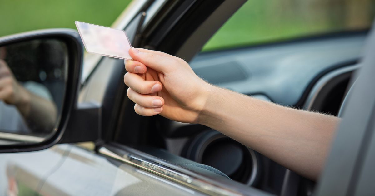 Motorista dentro de um carro estende a mão pela janela segurando um documento de licenciamento, com o retrovisor externo e parte do painel do veículo visíveis. A imagem representa a apresentação do CRLV durante uma fiscalização de trânsito.